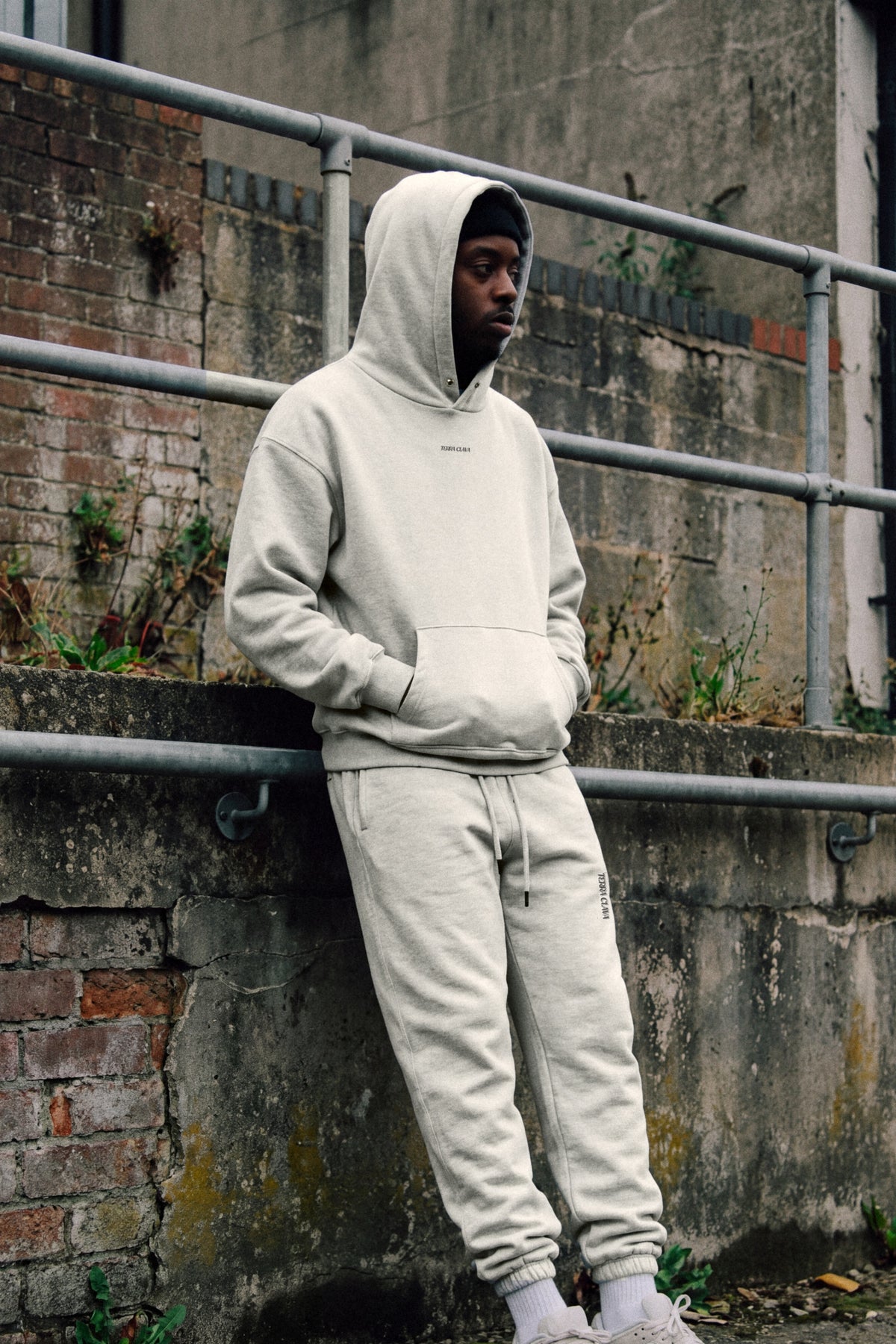 Alma Snap Collar Hoodie and Alma Sweatpants in Heather Gray, worn, embroidered logotype on chest and thigh, leaning on a railing