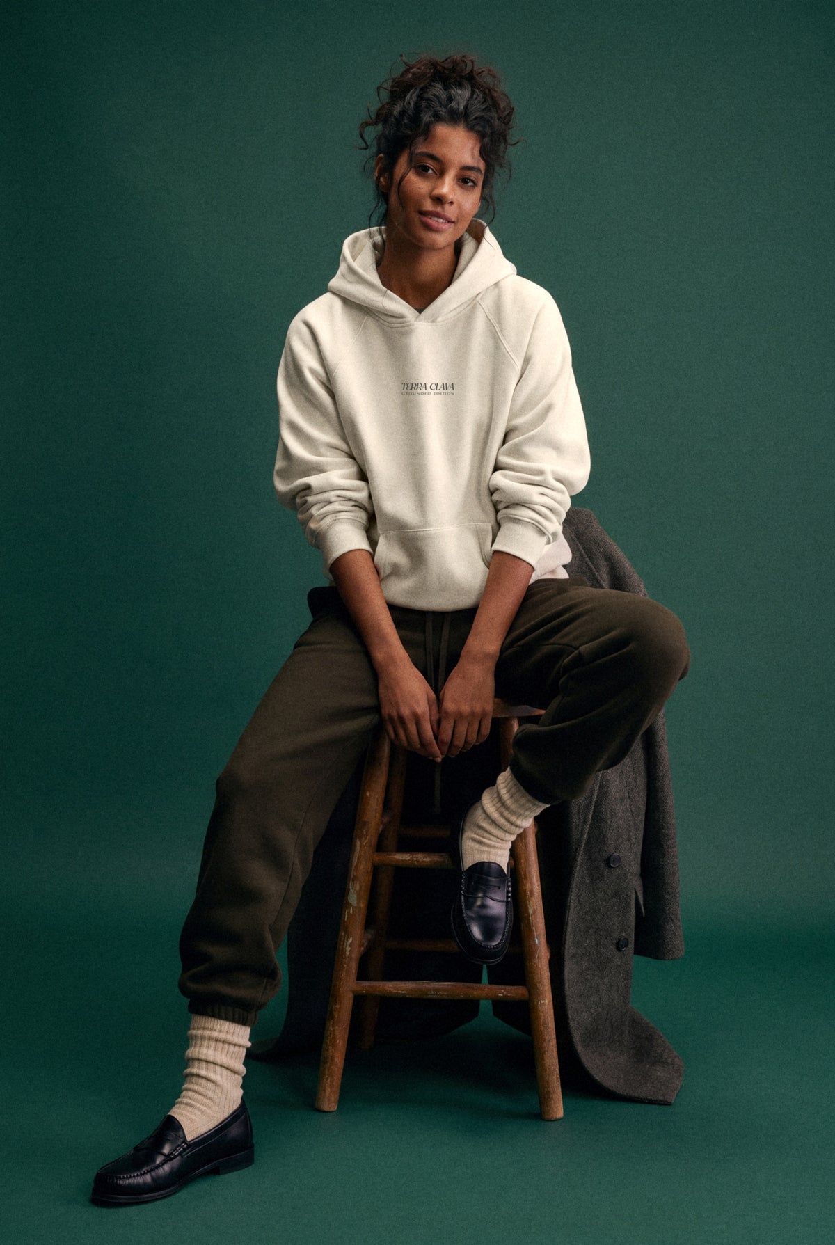 Grounded Edition Hoodie - Light in Rice Apricot, worn, chest print, seated on a wooden stool in a green studio